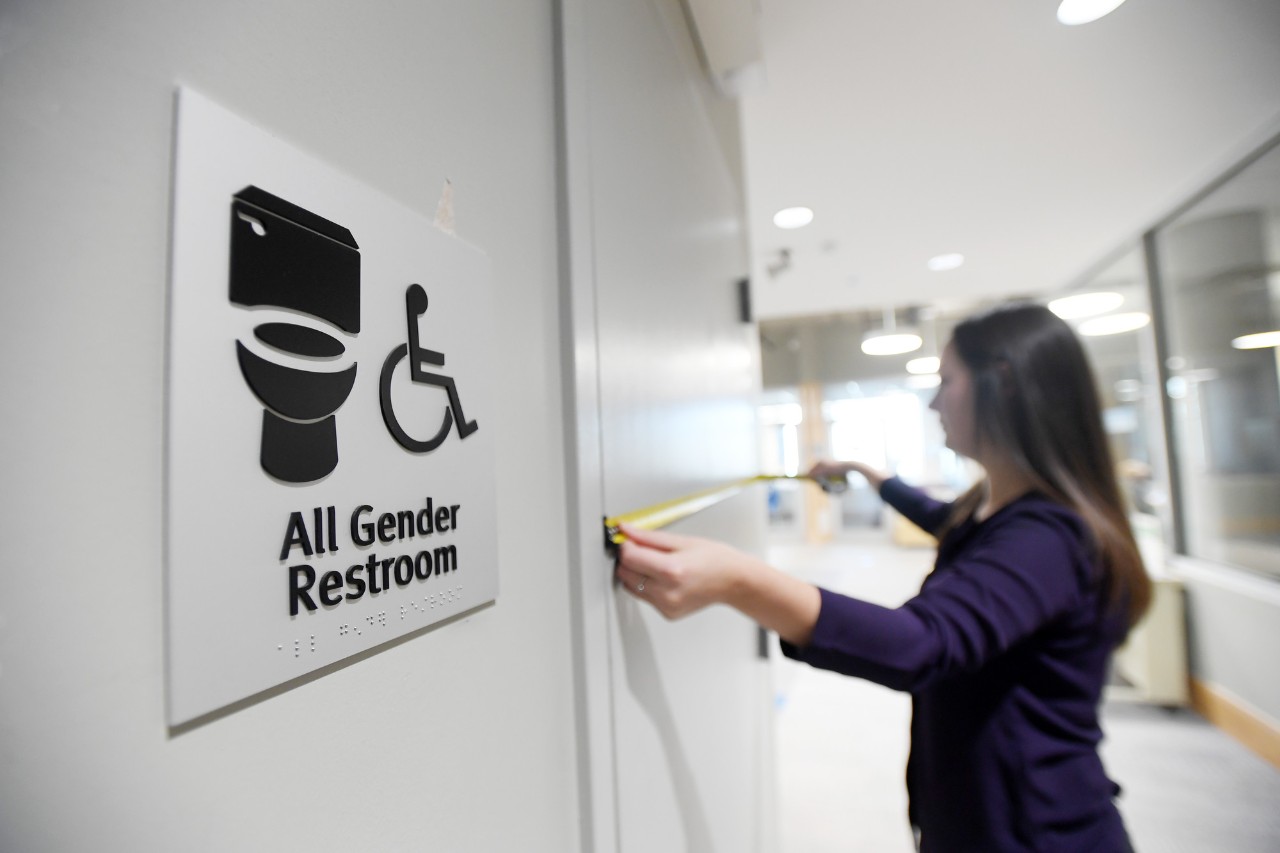 Person measures door next to all gender restroom sign.
